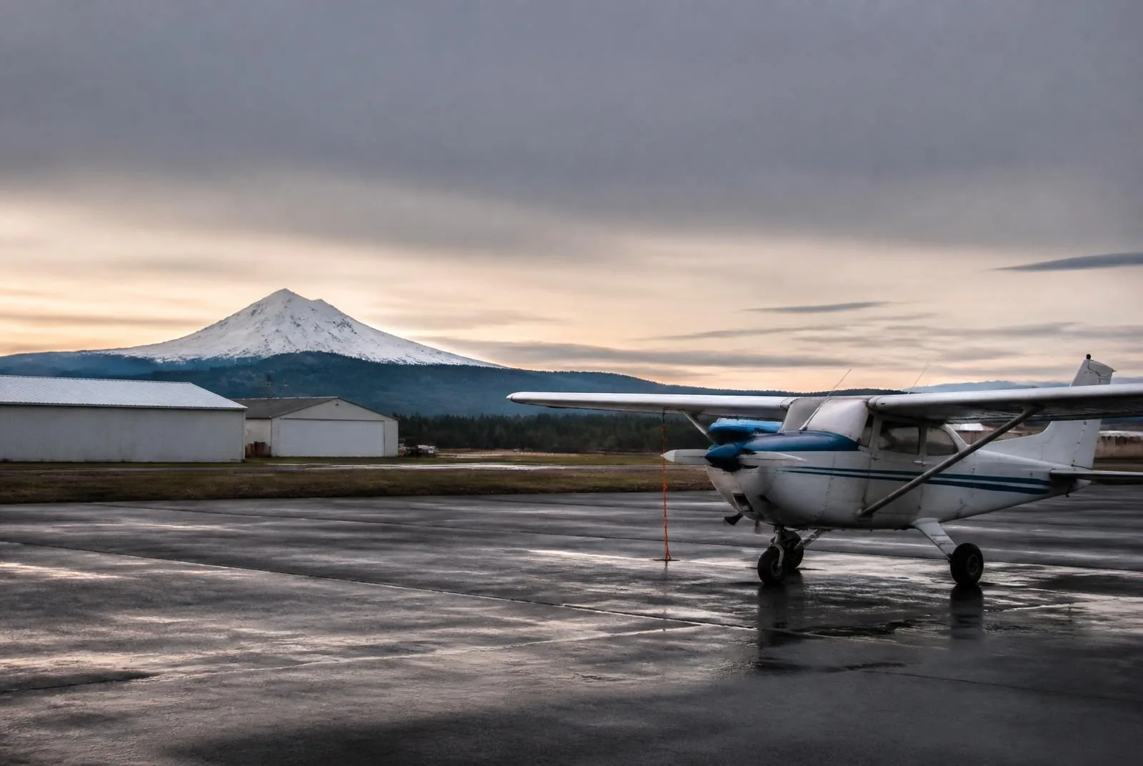 A single-engine light aircraft parked on a wet rural airfield ramp in Oregon with a snow-capped Cascade peak in the distance.